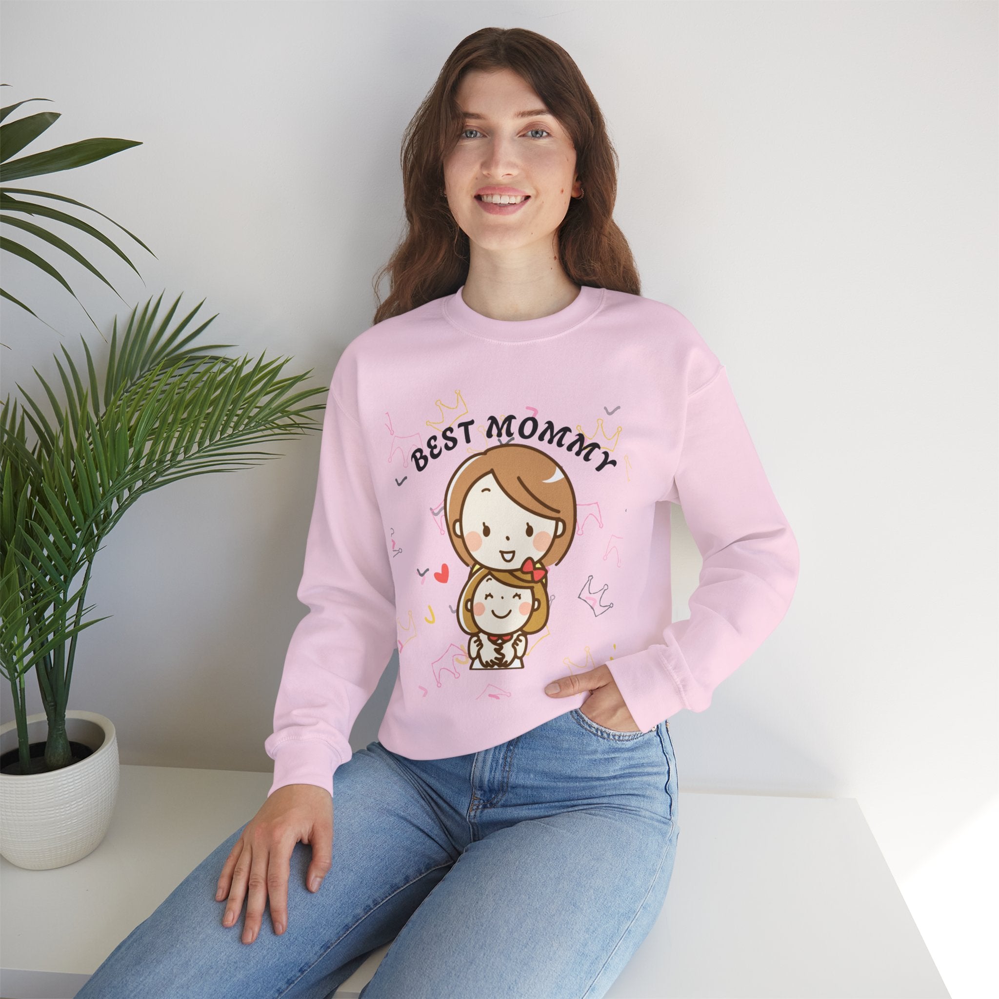 Woman wearing a Light pink sweatshirt with 'Best Mommy' text and cartoon character, sitting on a white surface with a plant in the background.