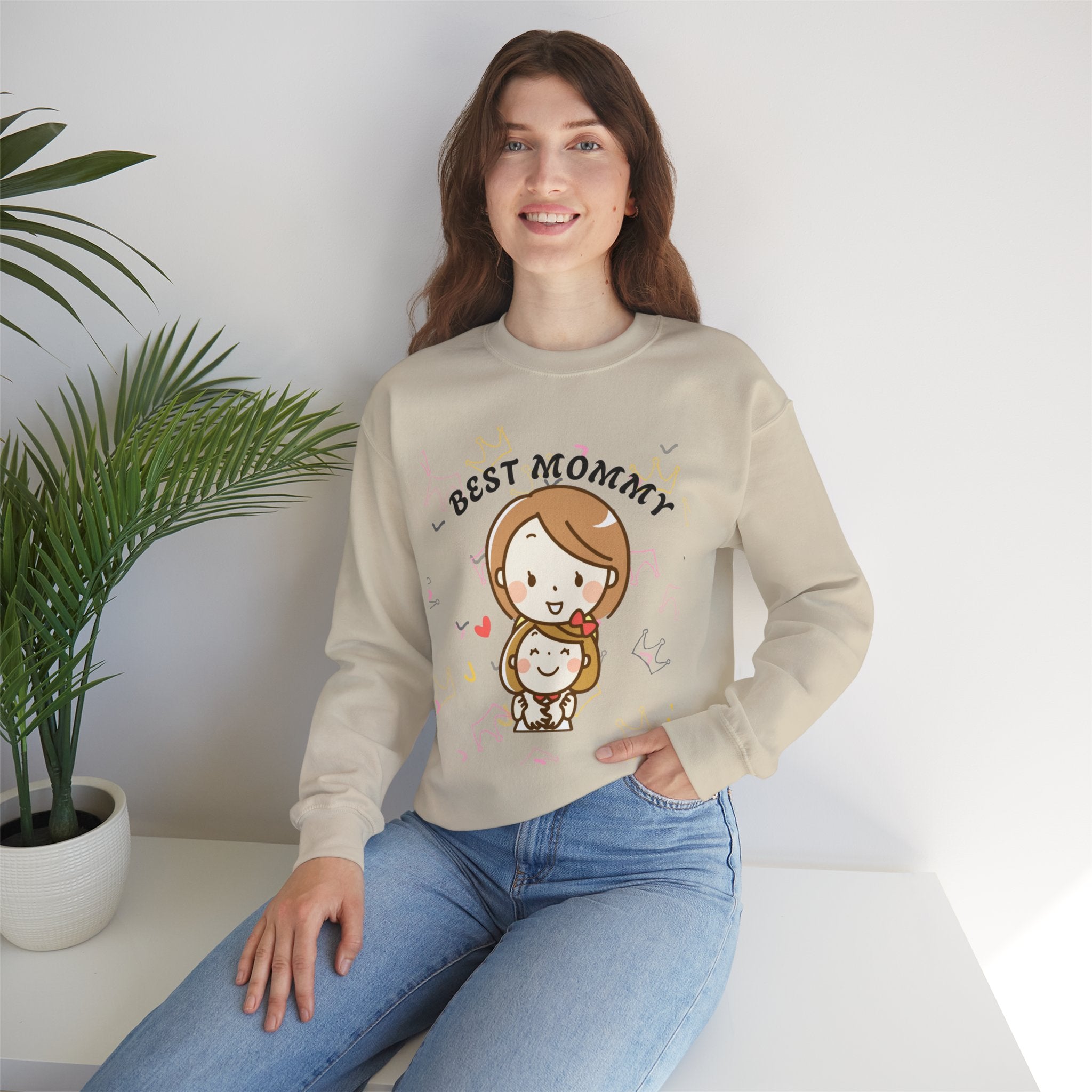 Woman wearing a Sand sweatshirt with 'BEST MOMMY' text and cartoon design, sitting on a white surface with a plant in the background.