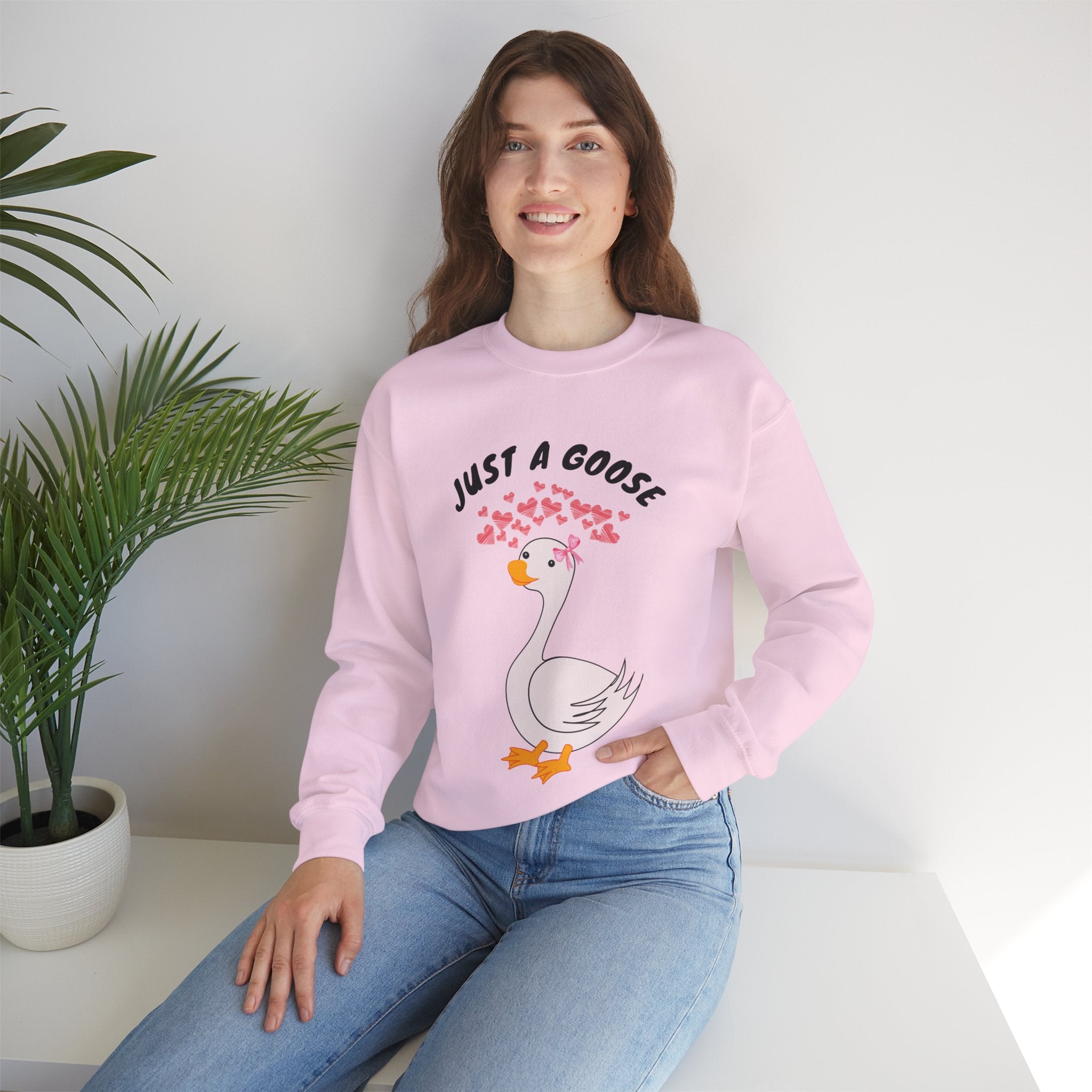 Person wearing a Light pink sweatshirt with a cartoon goose design and text, sitting on a white surface with a plant in the background.