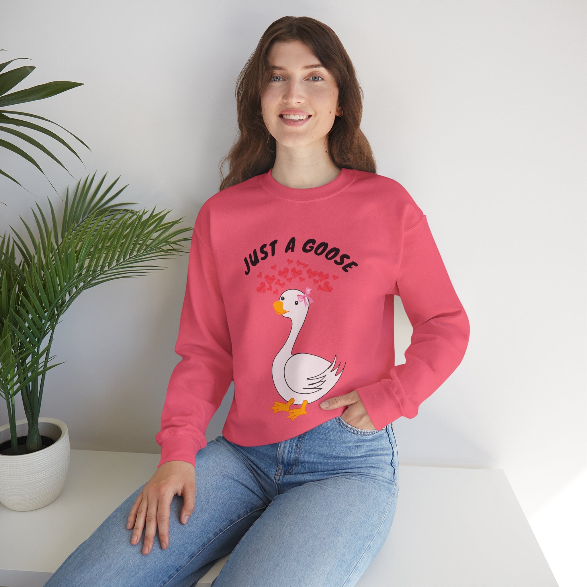 Person wearing a Safety pink sweatshirt with a cartoon goose and text, sitting on a white surface with a plant in the background.