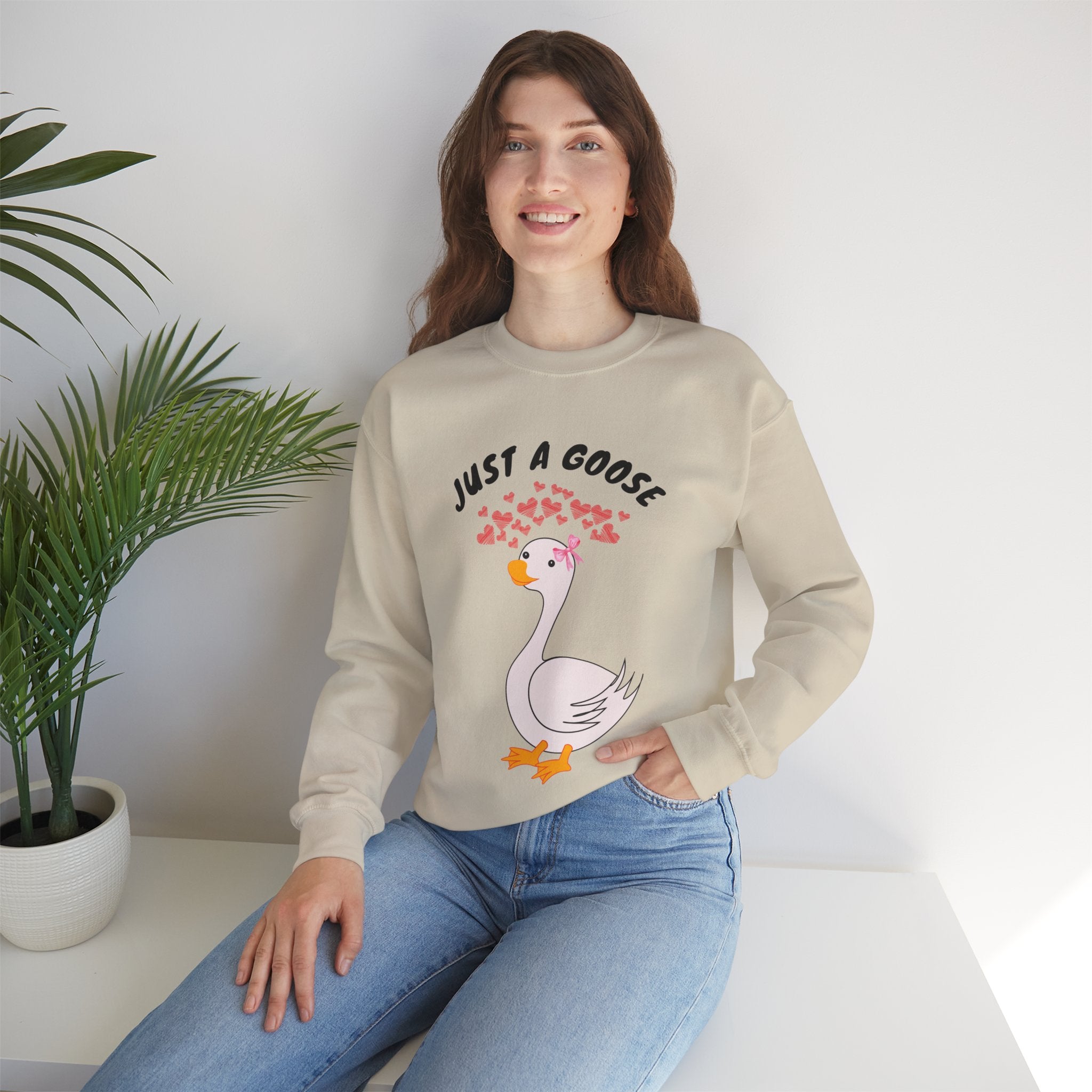 Woman wearing a Sand sweatshirt with a cartoon goose design and text, sitting on a white surface with a plant in the background.