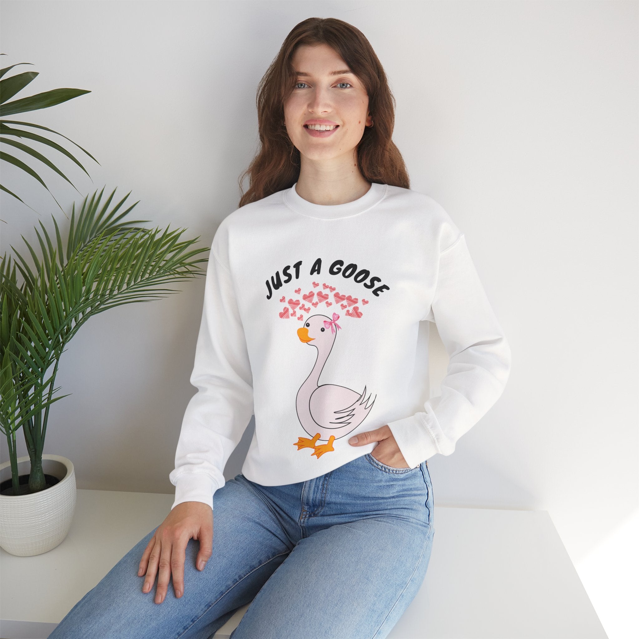 Woman wearing a white sweatshirt with a cartoon goose design and text, sitting on a white surface with a plant in the background.