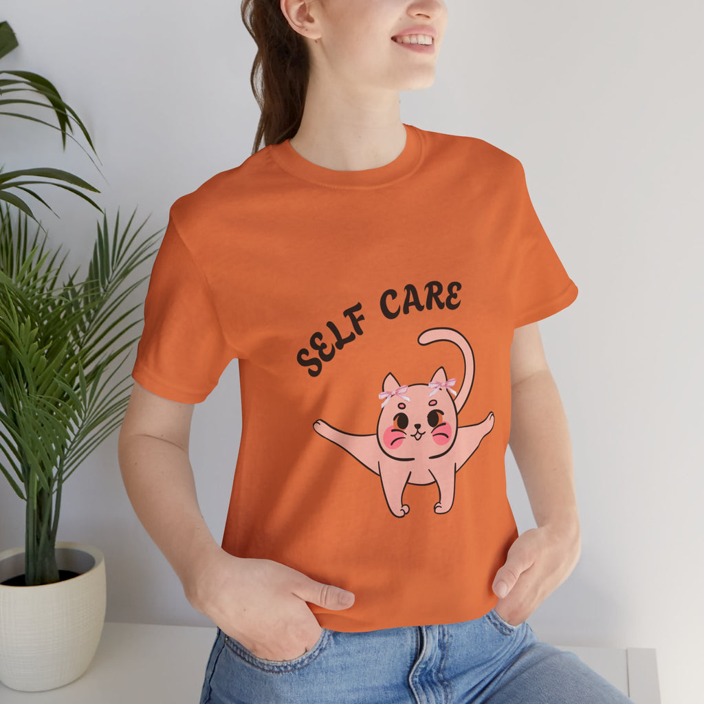 Person wearing a Burnt orange t-shirt with a cartoon cat and 'SELF CARE' text, sitting indoors.