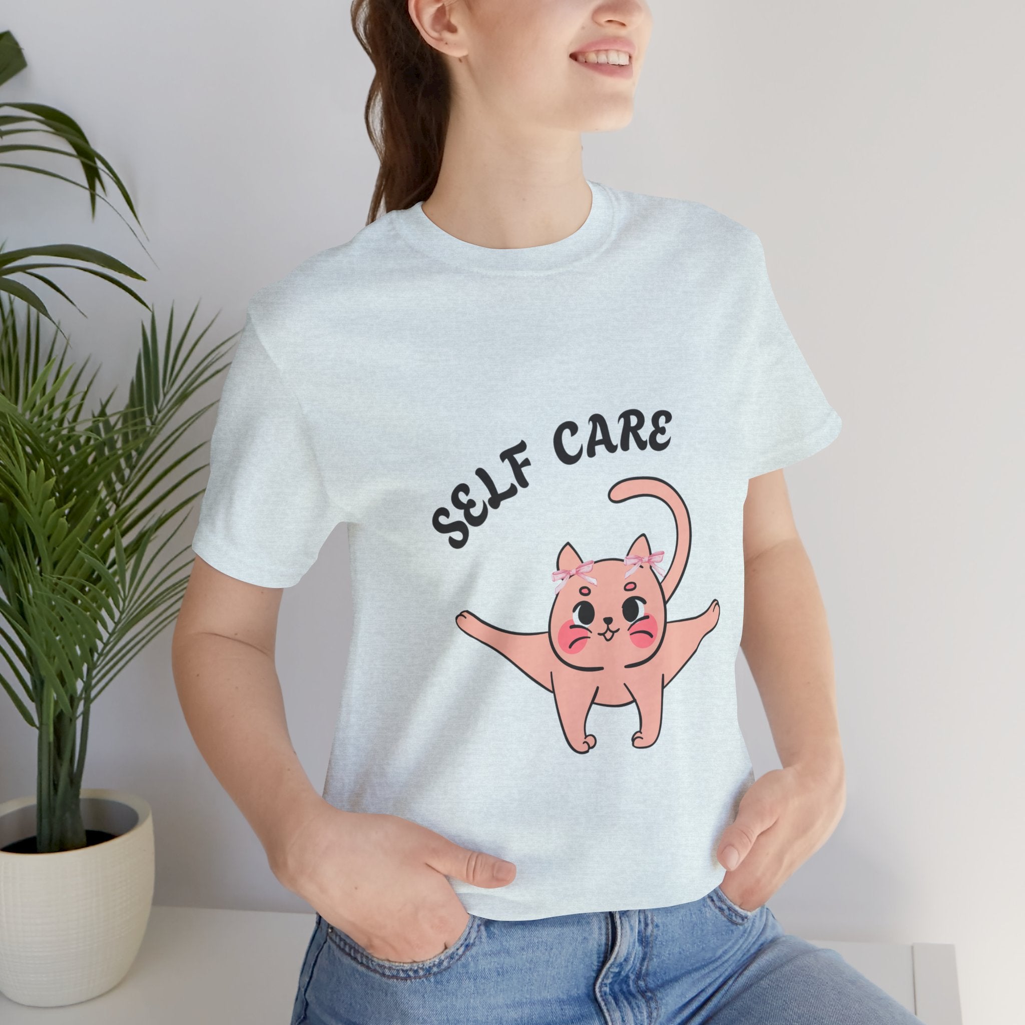 Person wearing a Heather Ice Blue t-shirt with a pink cat illustration and 'SELF CARE' text.