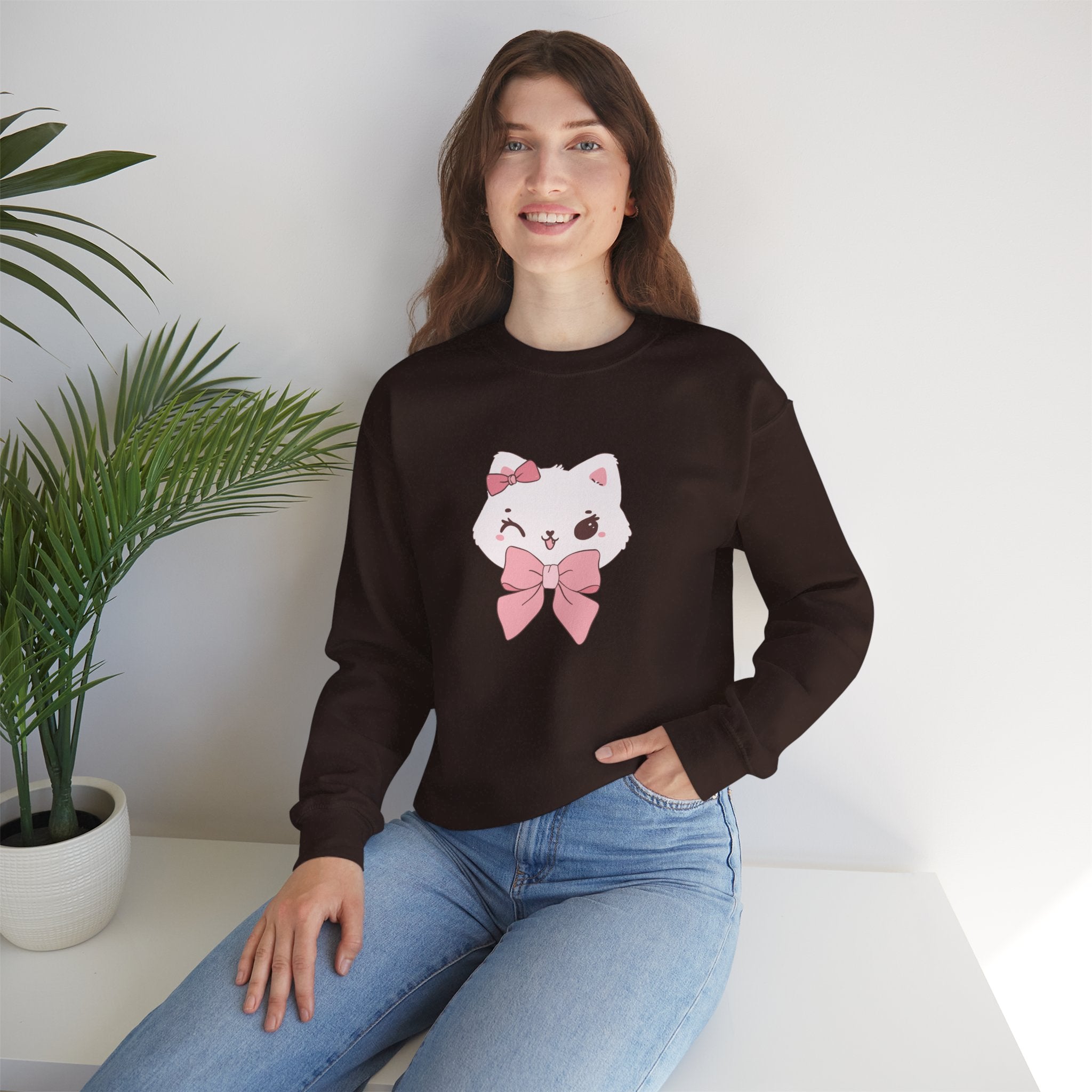 Woman wearing a Dark Chocolate sweatshirt with a white cat design and pink bow, sitting on a white surface with a plant in the background.
