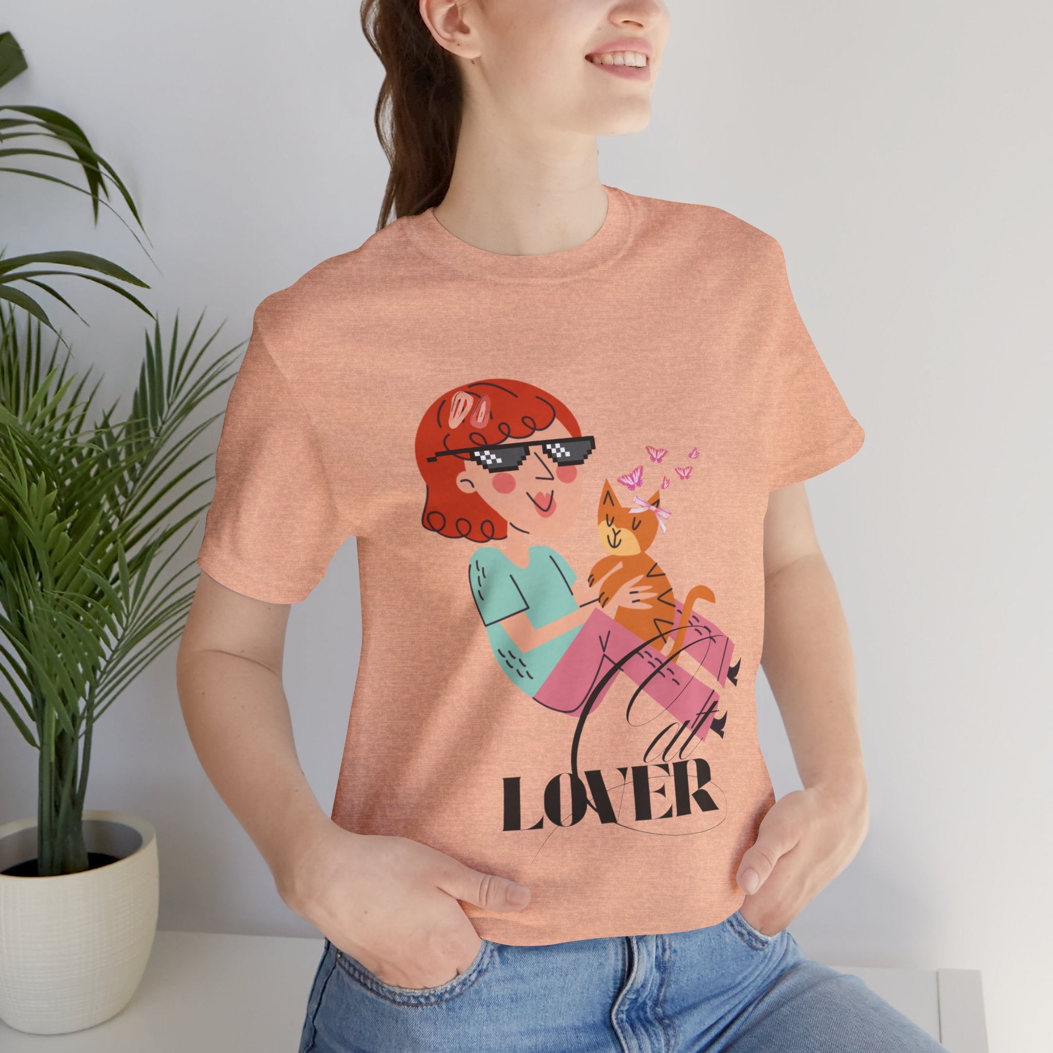 Person wearing a Heather peach t-shirt with a graphic design of a woman and cat, with 'Cat LOVER' text, in a minimal indoor setting.