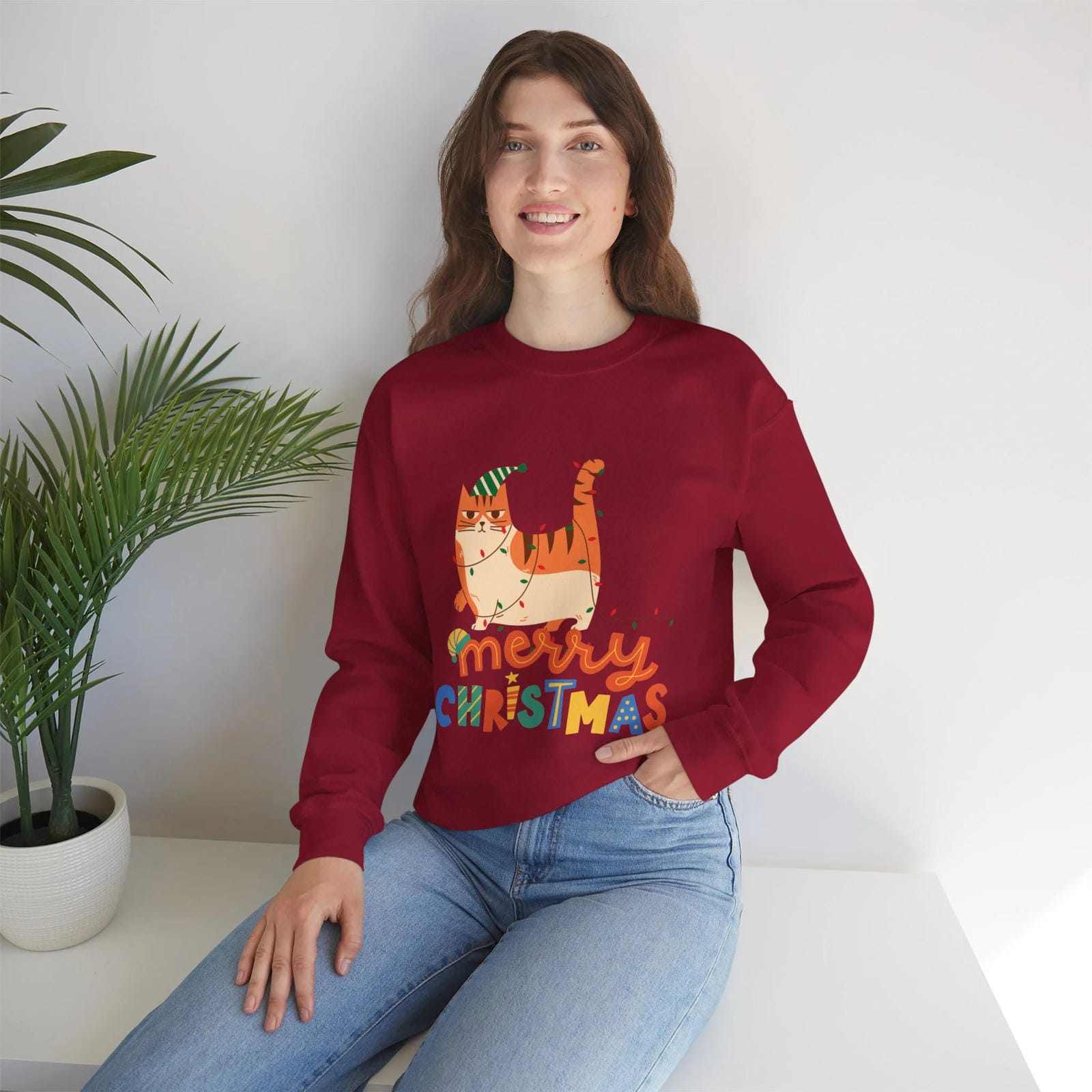 Person wearing a Cherry red 'Merry Christmas' sweater with a cat design, sitting on a white surface with a plant in the background.