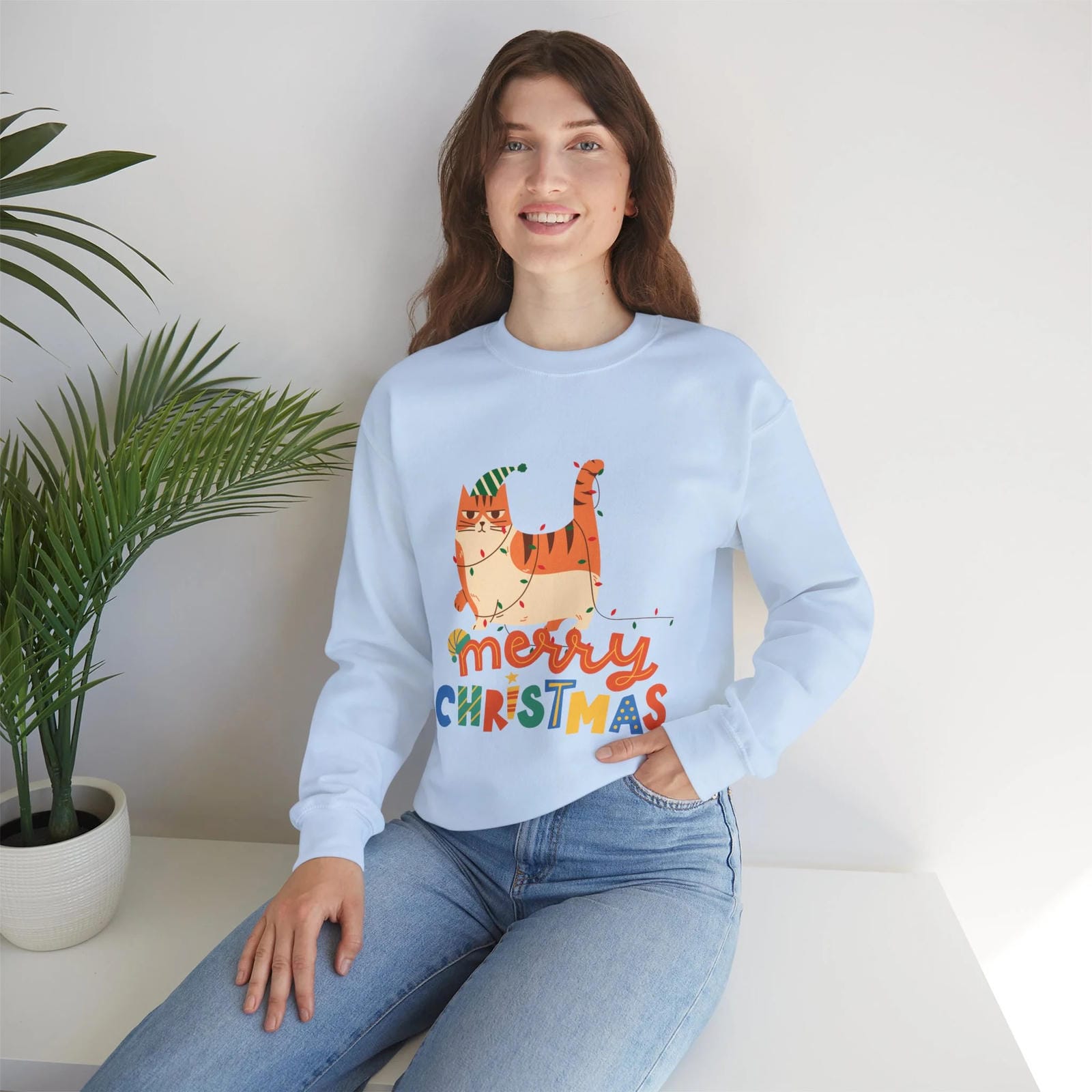 Woman wearing a light blue sweatshirt with a Christmas design, sitting on a white surface with a plant in the background.