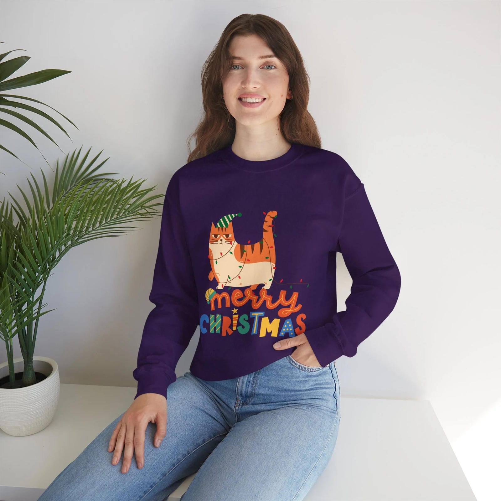 Person wearing a purple 'Merry Christmas' sweater with a cat design, sitting on a white surface with a plant in the background.