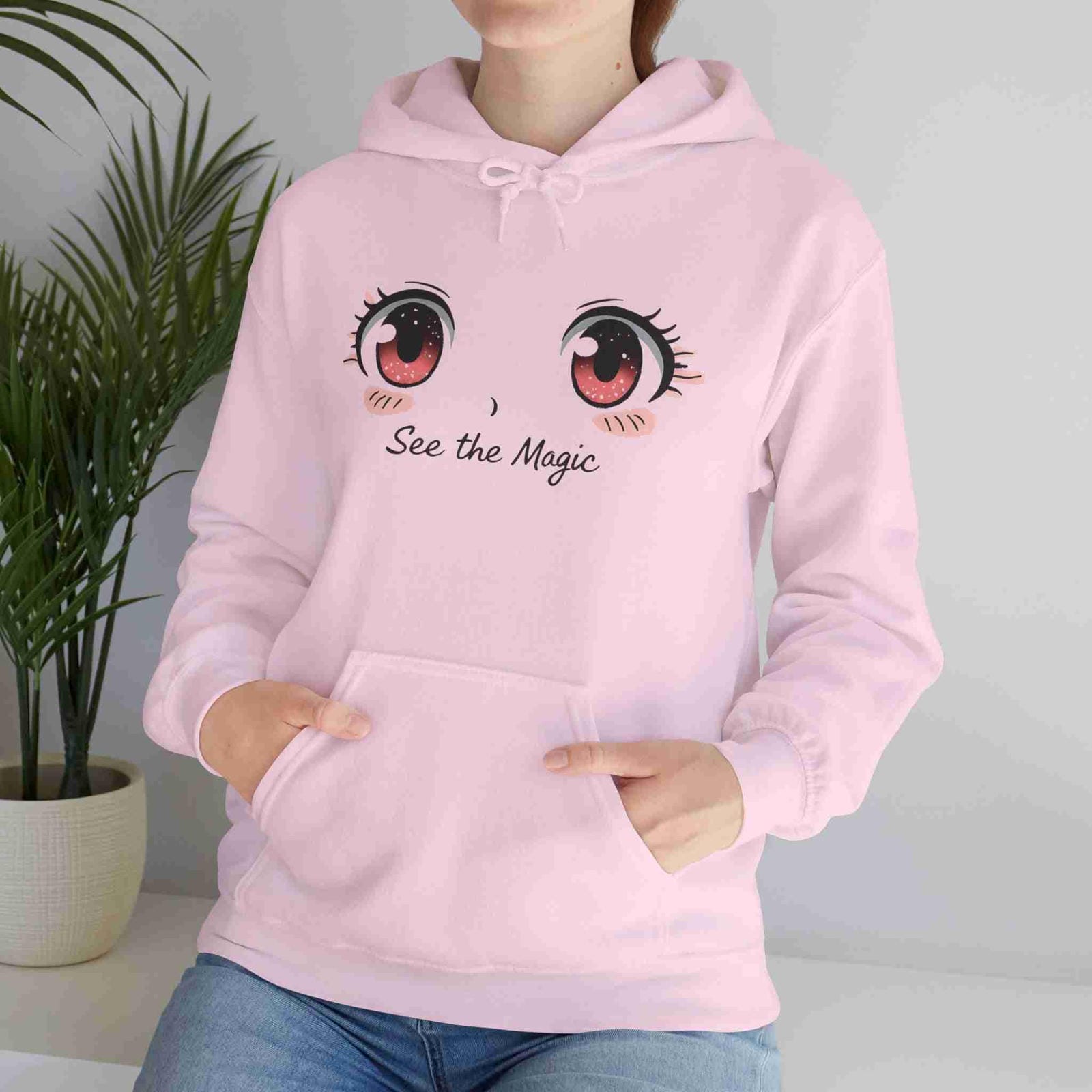 Person wearing a light pink hoodie with cartoon eyes and 'See the Magic' text, standing next to a plant.